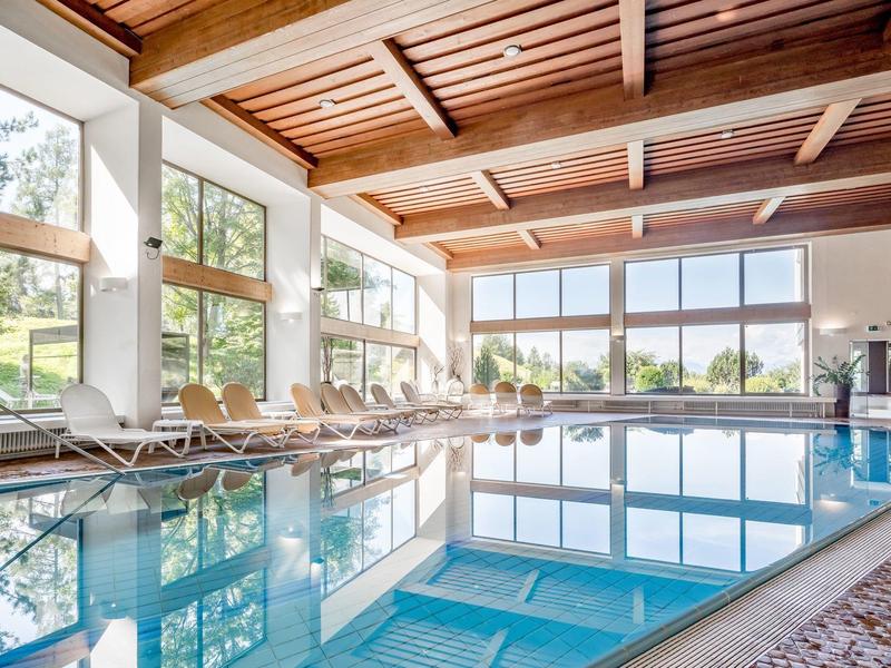 Helles Hallenbad mit großen Fenstern, Holzdecke, Liegestühlen und klarem blauen Wasser.