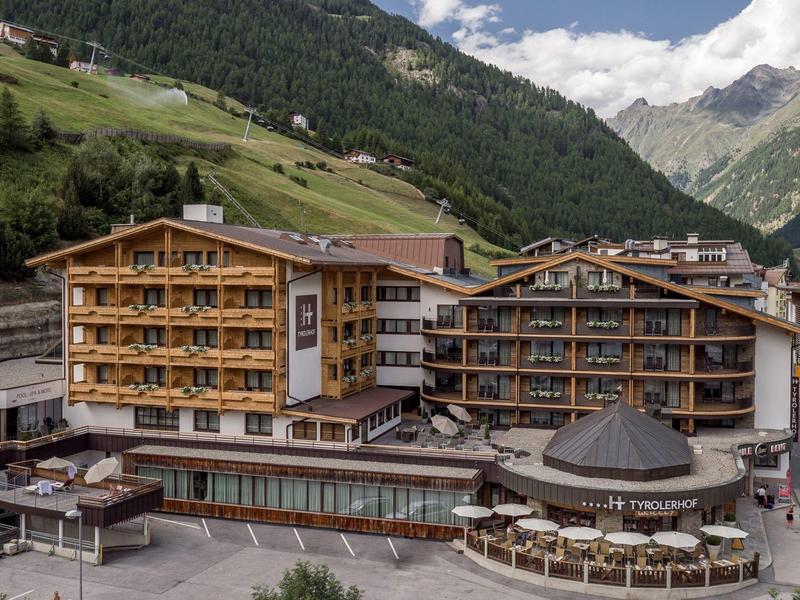 Duży hotel z drewnianą elewacją w alpejskim krajobrazie górskim pod niebieskim niebem.