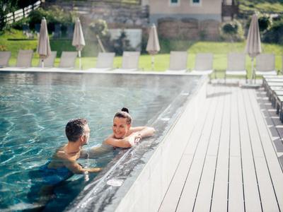 Two people relax in a heated outdoor pool next to a modern wooden deck with lounge chairs.