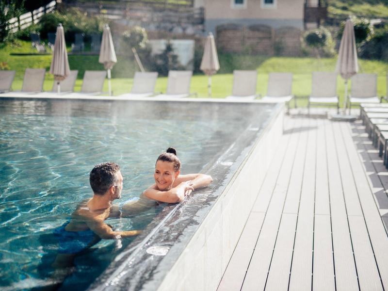 Two people relax in a heated outdoor pool next to a modern wooden deck with lounge chairs.