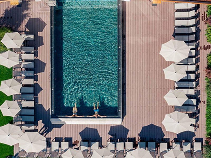 Aerial view of a rectangular pool with white lounge chairs and umbrellas on a terrace beside green lawns.