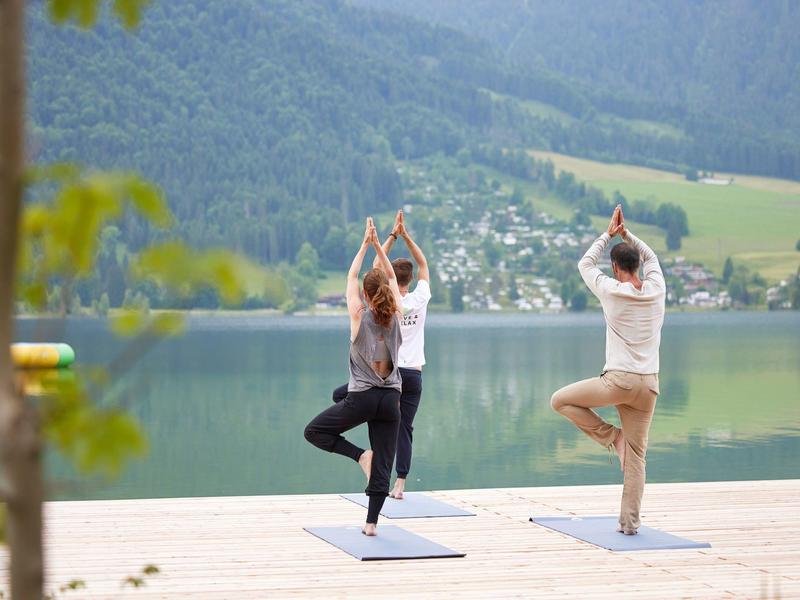 Three people practicing yoga on a wooden platform by a lake with forested mountains in the background.