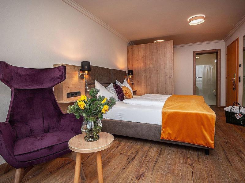 Gezellige hotelkamer met tweepersoonsbed, paarse stoel, houten vloer en bloemen op een klein tafel.