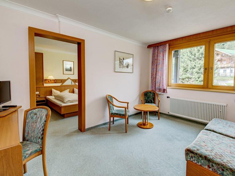 Helles Wohnzimmer mit Teppich, Sitzmöbeln, Fernseher und Blick ins Schlafzimmer mit Doppelbett.