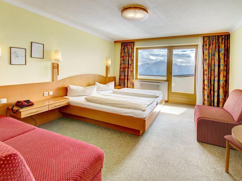 Helles Hotelzimmer mit Doppelbett, Sofa, Tisch, Nachttischlampen und Balkon mit Bergblick.