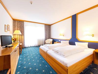 Helles Hotelzimmer mit Doppelbett, blauem Teppich, Fernseher und Fenster mit weißen Vorhängen.