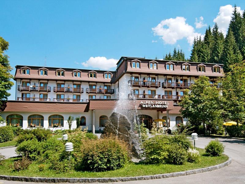 Großes Hotel mit mehrstöckigem Bau, Springbrunnen und grünen Büschen vor blauem Himmel.