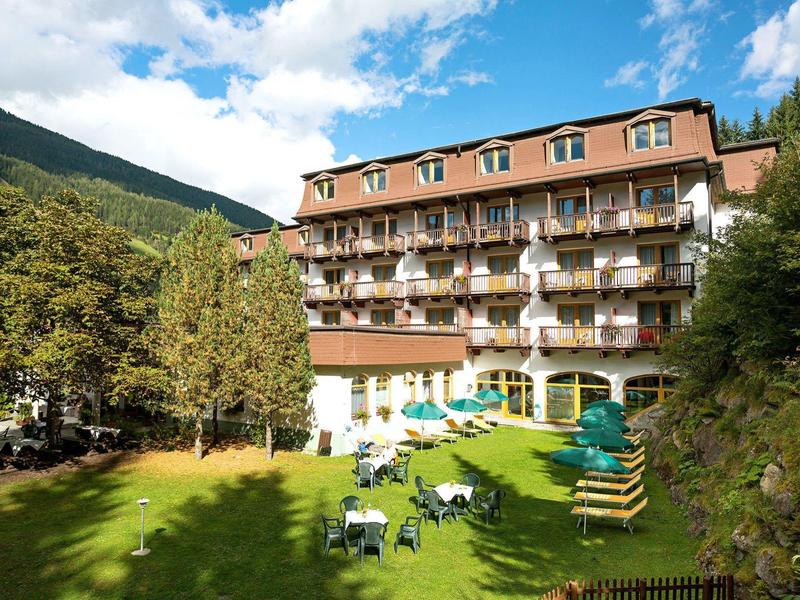 Großes Hotelgebäude mit Balkonen, umgeben von grüner Wiese, Bäumen und Bergen im Hintergrund.