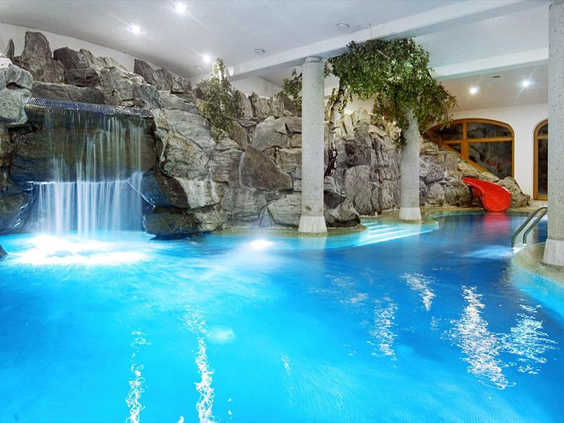 Innenpool mit Wasserfall, Felsenwand, heller Beleuchtung und roten Wasserrutschen.