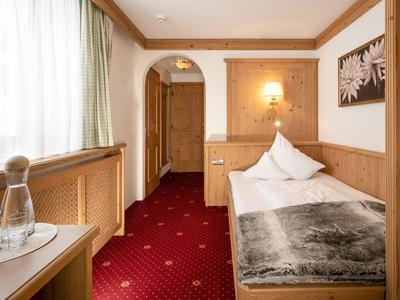 Gezellige eenpersoonskamer met houten meubels, rood tapijt en groot raam met gordijnen.