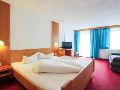Gemütliches Hotelzimmer mit Doppelbett, rotem Teppich, Sofa, TV und blauen Vorhängen am Fenster.