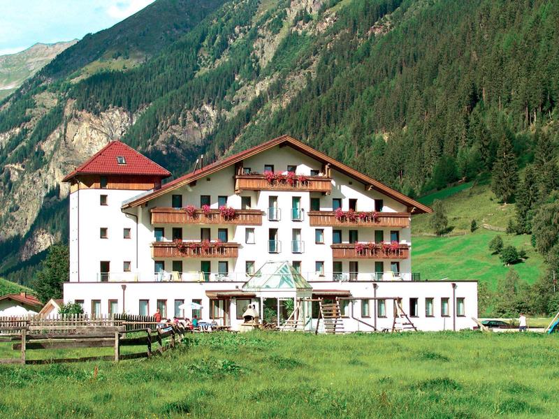 Großes Hotelgebäude vor bewaldeten Bergen und grüner Wiese im Tal.
