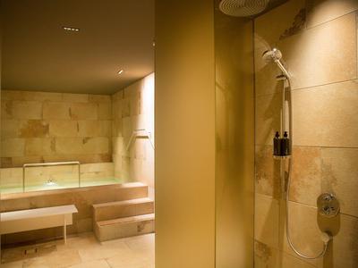 Área de bienestar cálida con bancos cómodos y ducha efecto lluvia en azulejos de piedra clara.