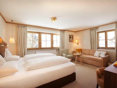 Habitación de hotel luminosa con dos camas, sofá, silla y grandes ventanas con cortinas.