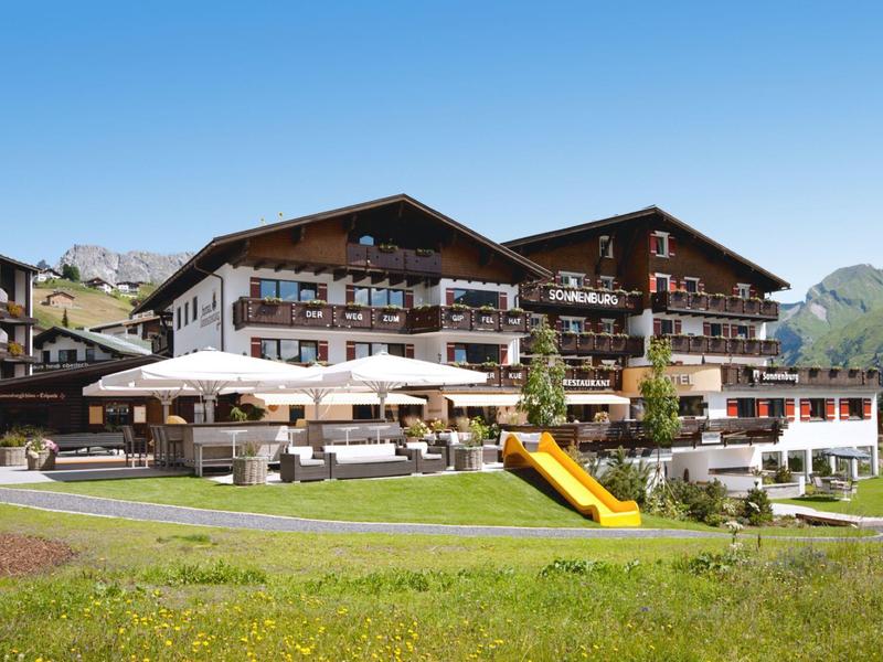 Gran hotel en las montañas con jardín, sombrillas y tobogán bajo un cielo despejado.