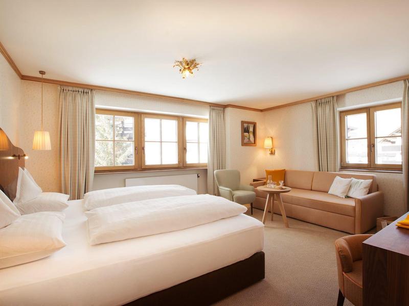 Habitación de hotel luminosa con dos camas, sofá, silla y grandes ventanas con cortinas.