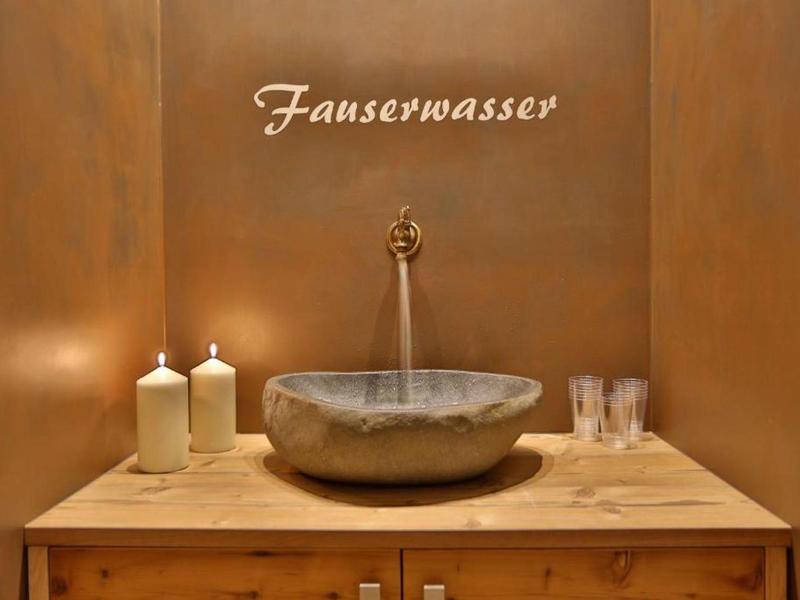 Rustikales Waschbecken auf Holzkommode mit zwei Kerzen und Gläsern vor braunem Hintergrund.