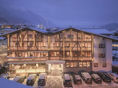 Beleuchtetes Berghotel mit Balkonen und geparkten Autos bei Dämmerung in den Alpen.