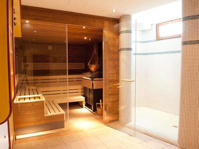 Moderne Sauna mit Holzverkleidung, Glaswand, Holzbänken und einem Ofen, heller Fliesenboden.