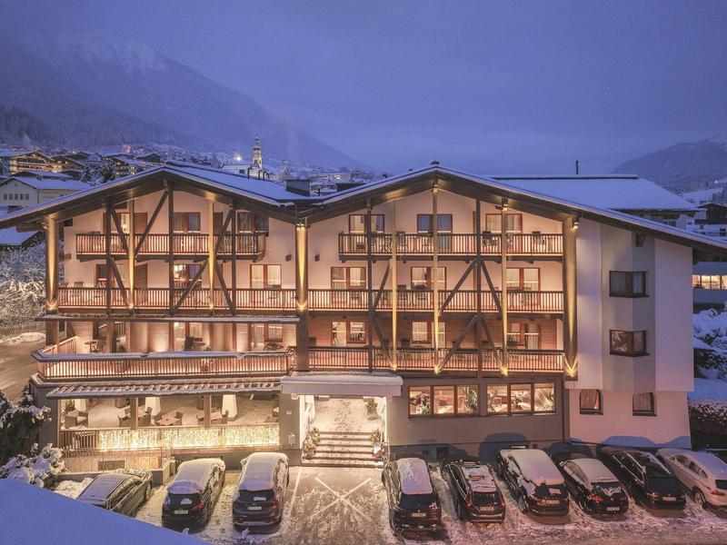 Beleuchtetes Berghotel mit Balkonen und geparkten Autos bei Dämmerung in den Alpen.