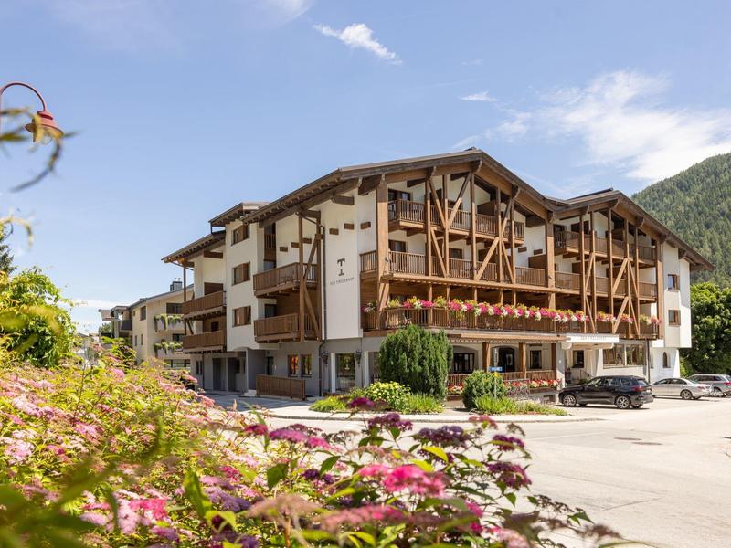 Großes Alpenhotel mit Holzfassade, Balkonblumen und blühendem Garten vor Bergkulisse.