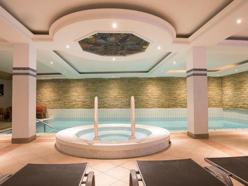 Helles Hallenbad mit runder Whirlwanne, Liegestühlen und dekorativer Decke in warmen Farben.