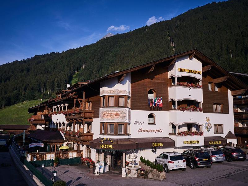 Traditionelles Hotel mit Balkon und Holzverkleidung vor bewaldetem Berg, Autos im Vordergrund.