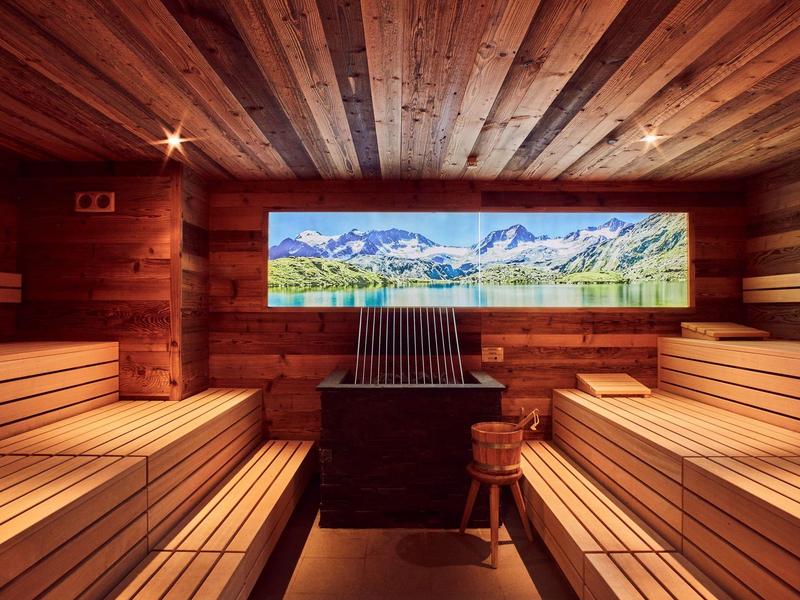Sauna mit hellem Holz, breiten Holzbänken und großem Panoramabild einer Berglandschaft.
