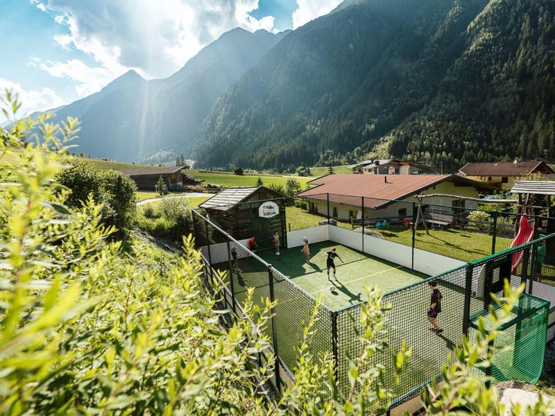 Ein Tennisplatz in einem grünen Tal mit Bergen und Häusern im Hintergrund, unter blauem Himmel.