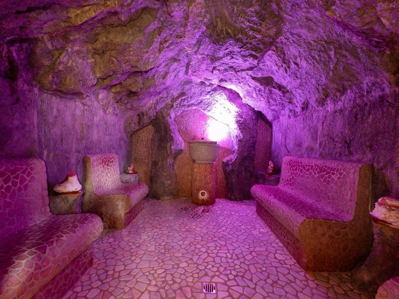 Gemütlicher, mit violettem Licht beleuchteter Raum in einer Höhle mit Sitzgelegenheiten und Steintisch.