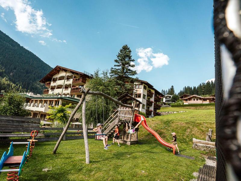 Spielplatz mit bunter Rutsche und Schaukel vor Holzhäusern in bergiger, grüner Landschaft.