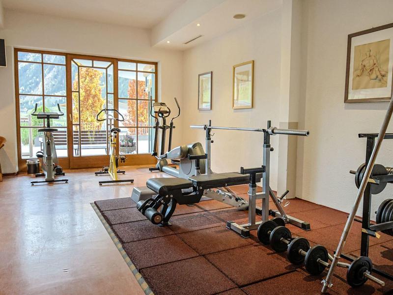 Fitnessraum mit Gewichthebestation, Geräten und großem Fenster mit Ausblick auf Bäume.