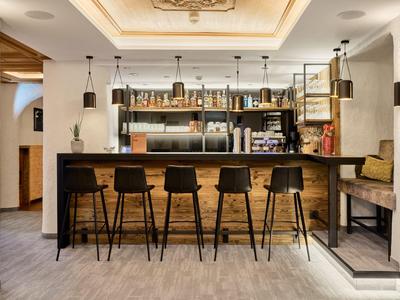 Área de bar moderna en hotel con mostrador de madera, taburetes y estantes con botellas y vasos.