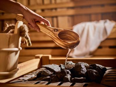 Hand gießt Wasser auf heiße Steine in einer Sauna mit Holzwänden und Holzeimer im Hintergrund.