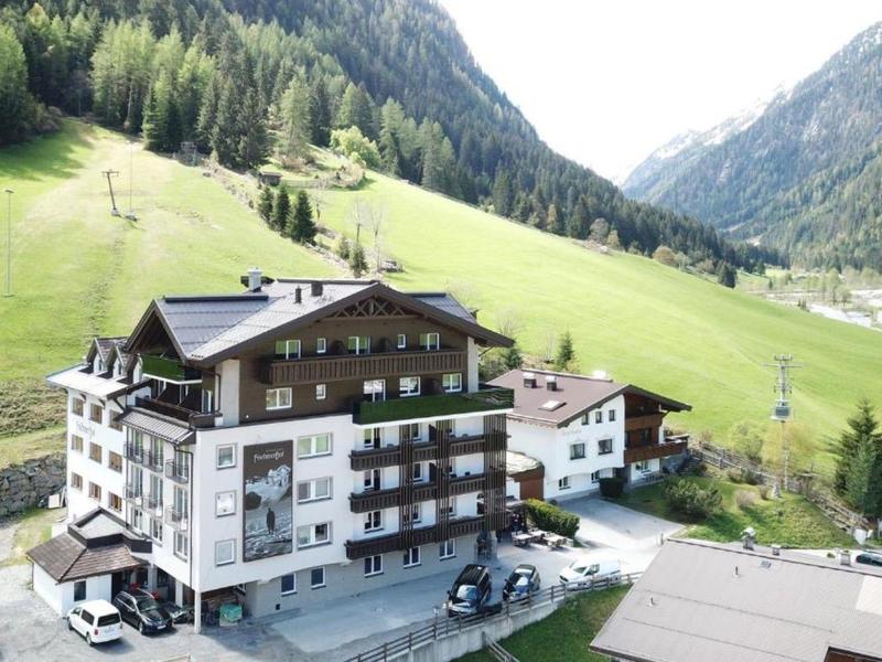 Mehrstöckiges Hotelgebäude in einem grünen Bergtal mit umliegenden Wiesen und bewaldeten Hängen.