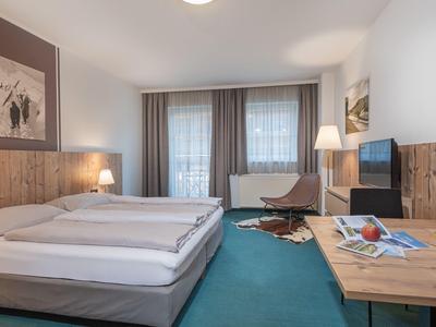 Modernes Hotelzimmer mit Doppelbett, türkisfarbenem Teppich und großem Fenster mit Gardinen.