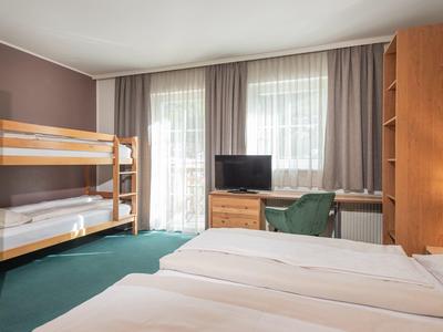 Schlafzimmer mit Etagenbett, Doppelbett, grünem Teppich und Holzmöbeln vor zwei großen Fenstern.