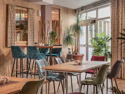 Gemütlicher Gastraum mit Holztischen, bunten Stühlen, Pflanzen und großen Fenstern.