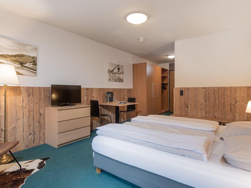 Doppelzimmer mit zwei Betten, Holzpaneelen, Schreibtisch, TV, blauer Teppich und Stehlampe.