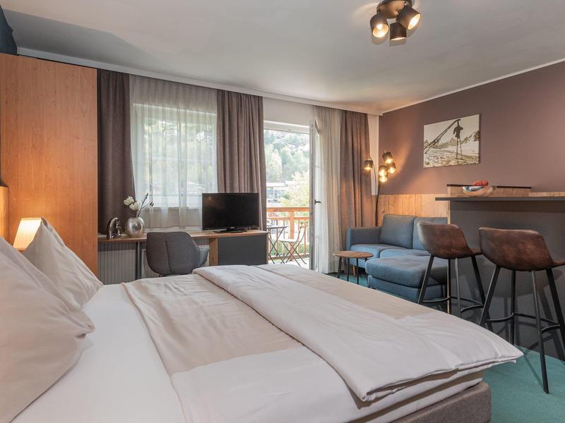 Modern eingerichtetes Hotelzimmer mit großem Bett, Sofa, Fernseher und Balkon mit Blick ins Grüne.