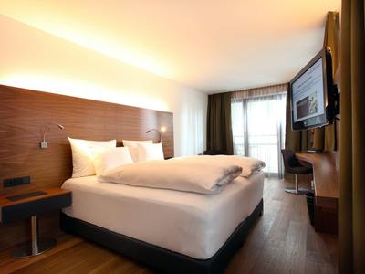 Helles Hotelzimmer mit großem Doppelbett, dunkelbraunem Holzhintergrund und TV an der Wand.