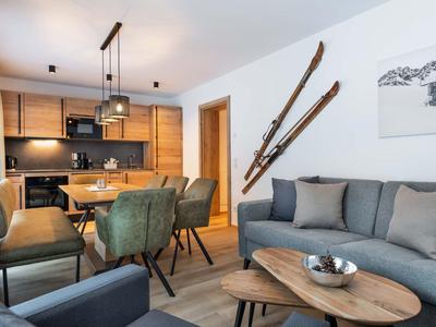 Wohnzimmer und Küche mit Holzmöbeln, grünen Stühlen, Ski an der Wand und grauer Couch.