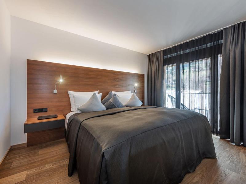 Modernes Schlafzimmer mit großem Bett, dunkler Tagesdecke, Holzwand und bodentiefen Fenstern.