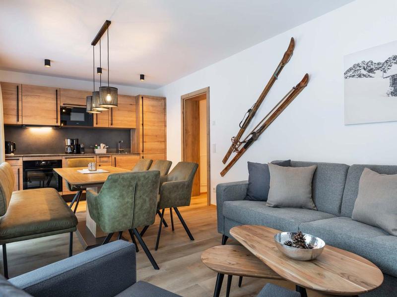 Wohnzimmer und Küche mit Holzmöbeln, grünen Stühlen, Ski an der Wand und grauer Couch.