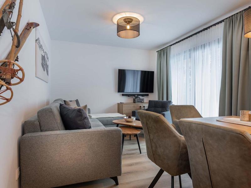 Gemütliches Wohnzimmer mit grauem Sofa, Esstisch, Stühlen, TV und großen Fenstern mit Vorhängen.