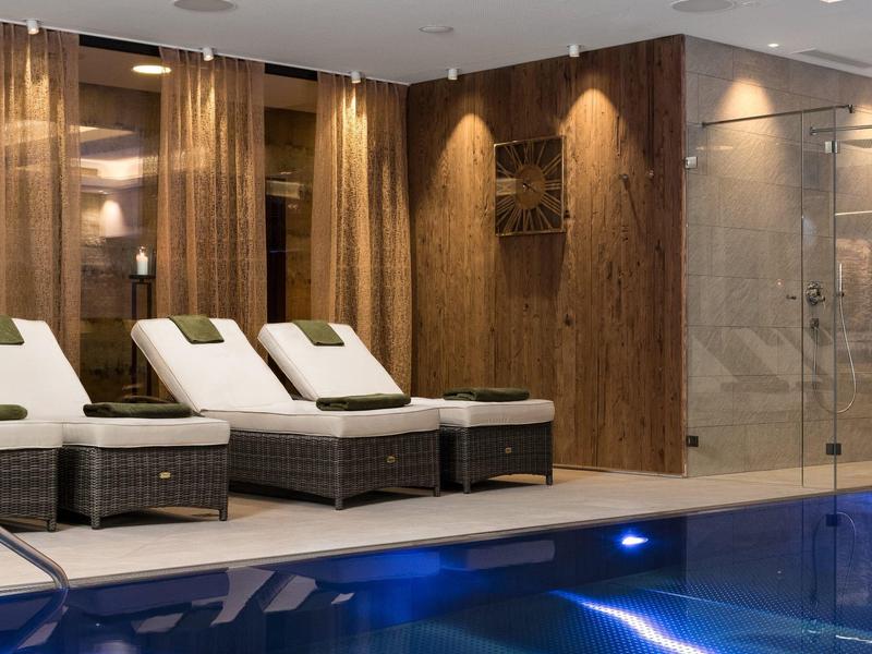 Innenraum mit blau beleuchtetem Schwimmbad und weißen Liegestühlen neben Holzwänden.