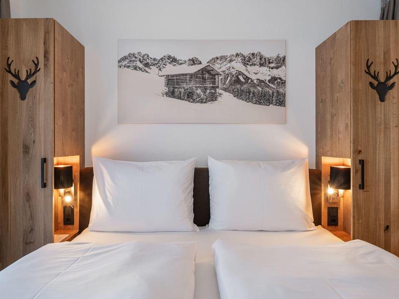 Doppelbett mit weißen Kissen und Bettwäsche, zwei Holzschränke und Bergbild an weißer Wand.