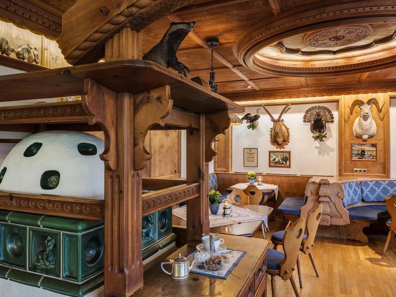 Gemütlicher Gastraum mit Holzmöbeln, grünem Kachelofen, antiken Trophäen und blauen Sitzbänken.