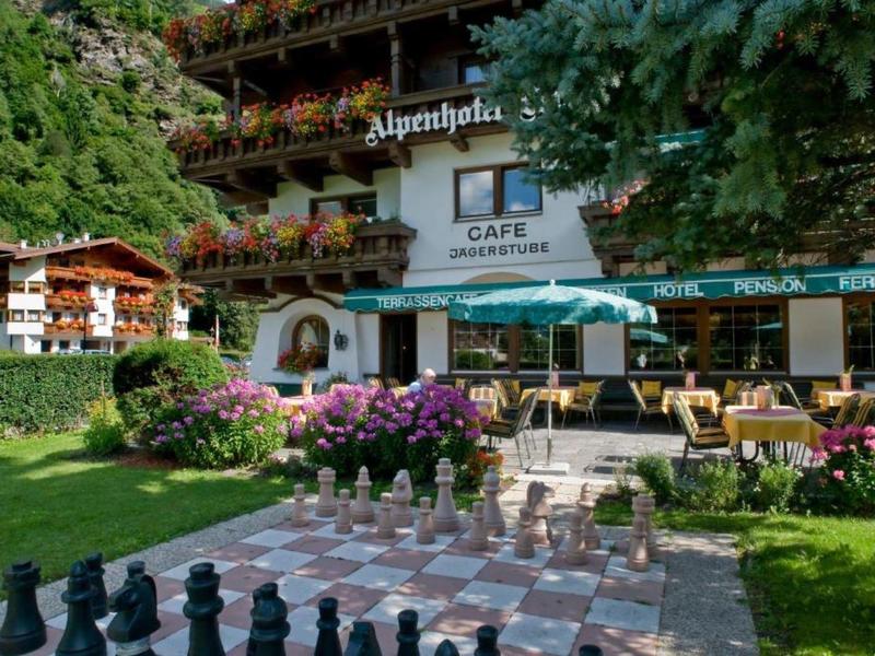 Großes Schachspiel vor traditionellem Alpenhaus mit Balkon und Außencafé im Grünen.