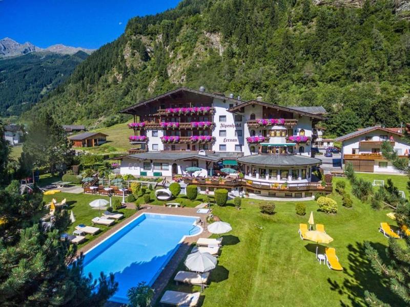 Großes weißes Alpenhotel mit violetten Blumentöpfen, großem Pool und grüner Wiese im Bergtal.